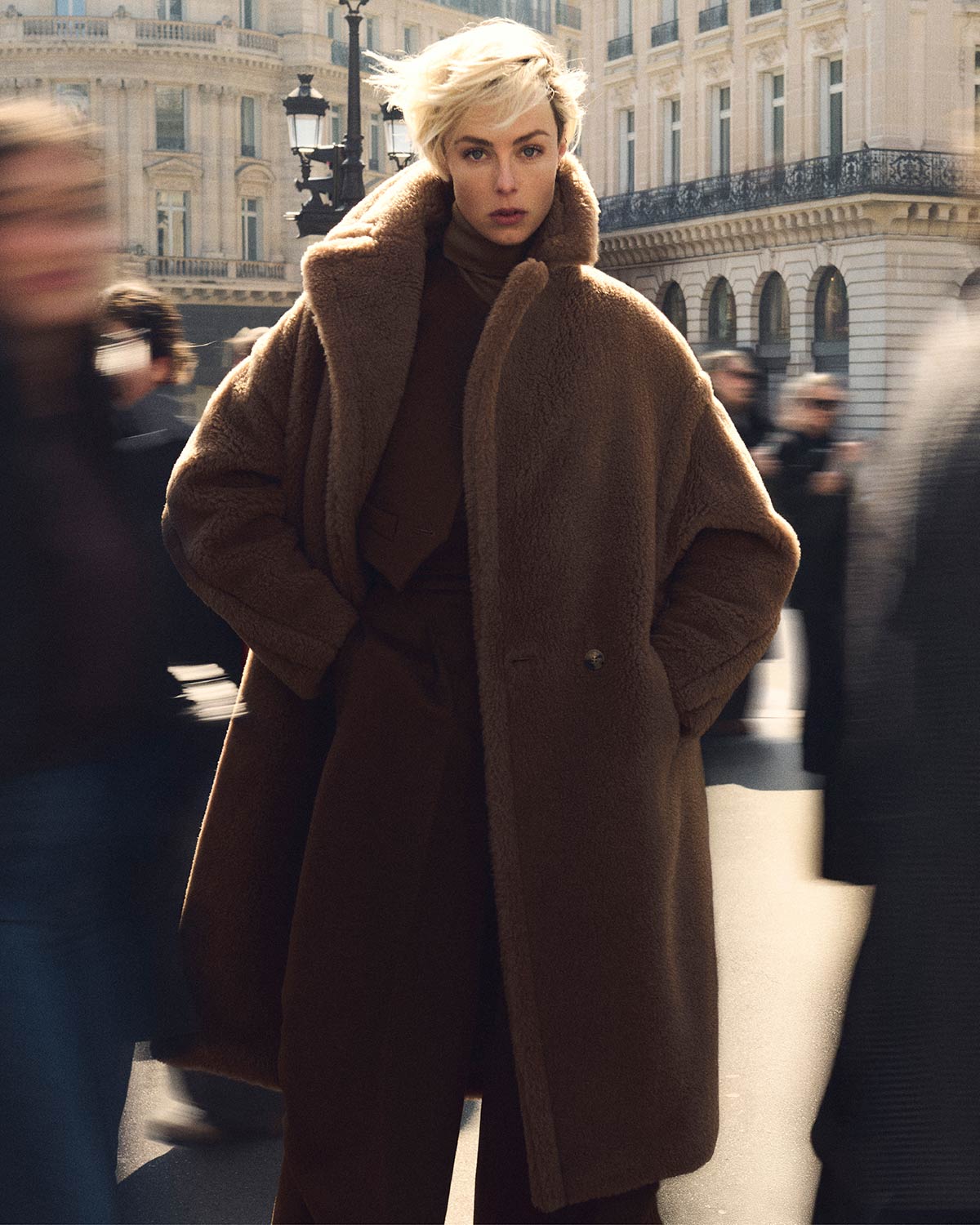 Model with Max Mara long Teddy Bear icon coat in camel colour, in a bustling urban environment.